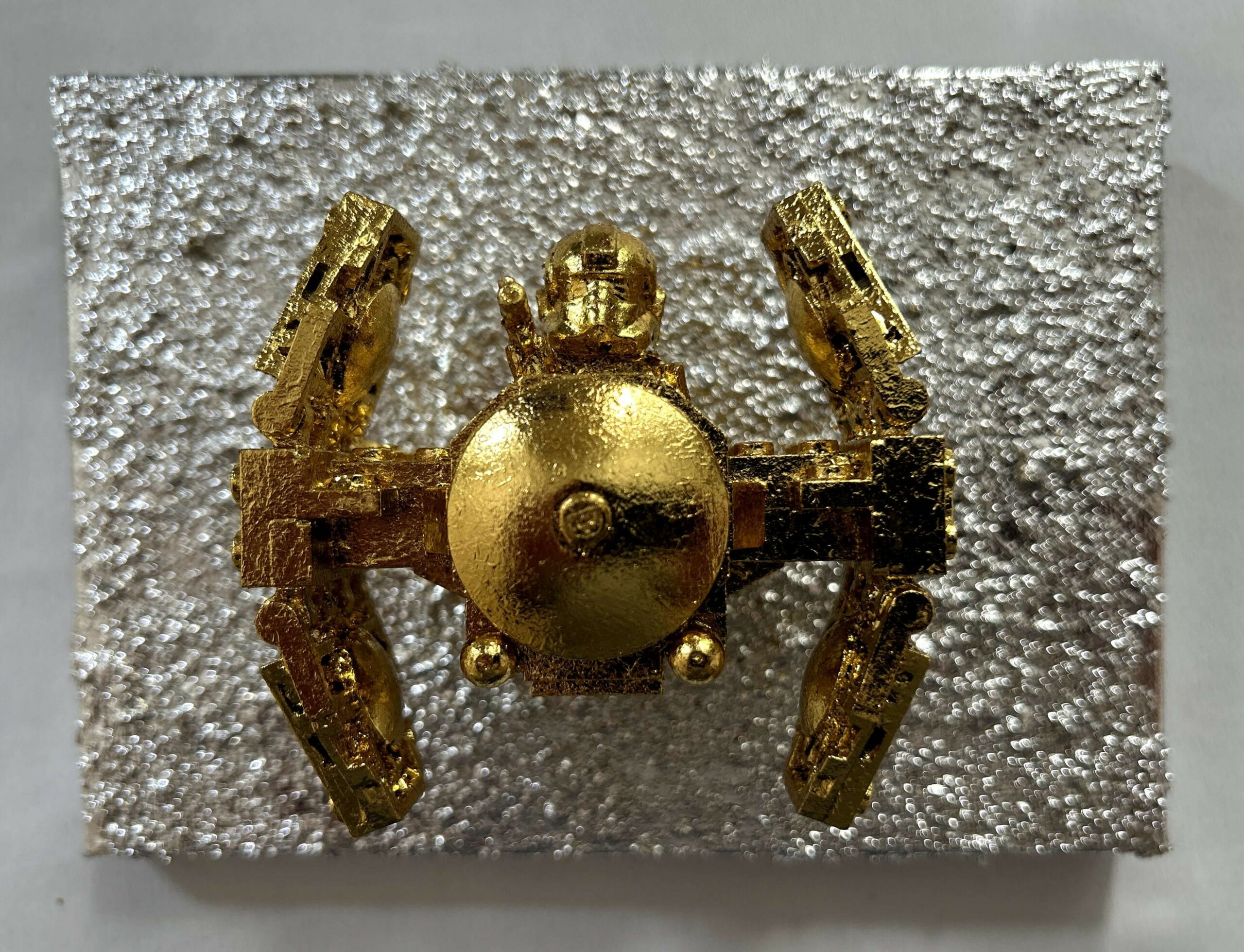 Seb Machten contemporary artwork with gold starfighter built from construction bricks projecting from textured silver surface