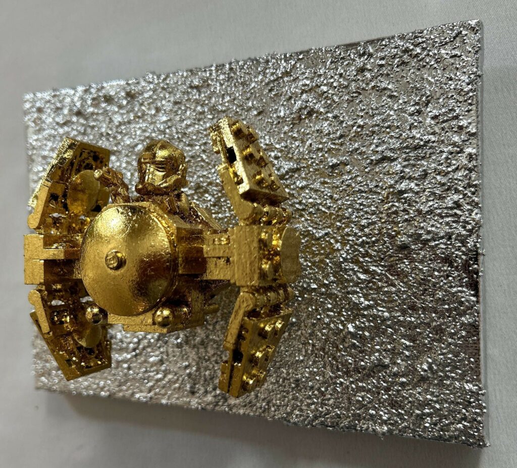 Seb Machten contemporary artwork with gold starfighter built from construction bricks projecting from textured silver surface