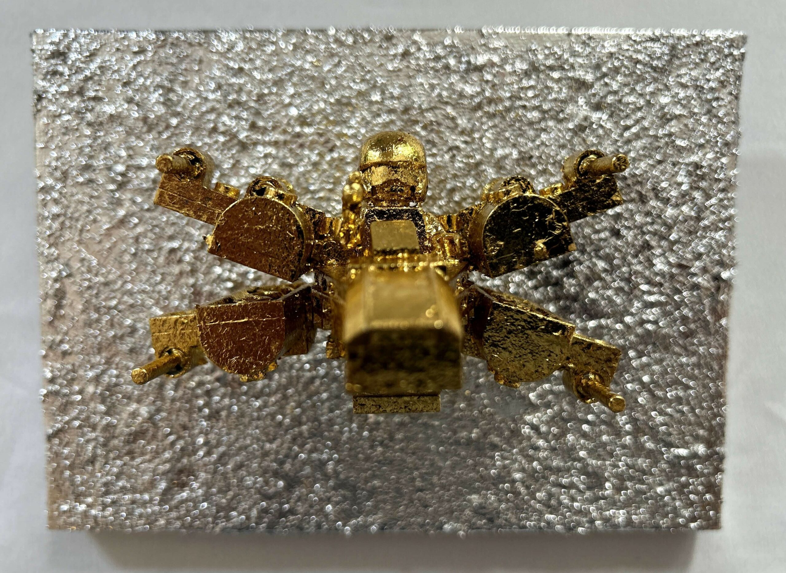 Seb Machten contemporary artwork with gold starfighter built from construction bricks projecting from textured silver surface