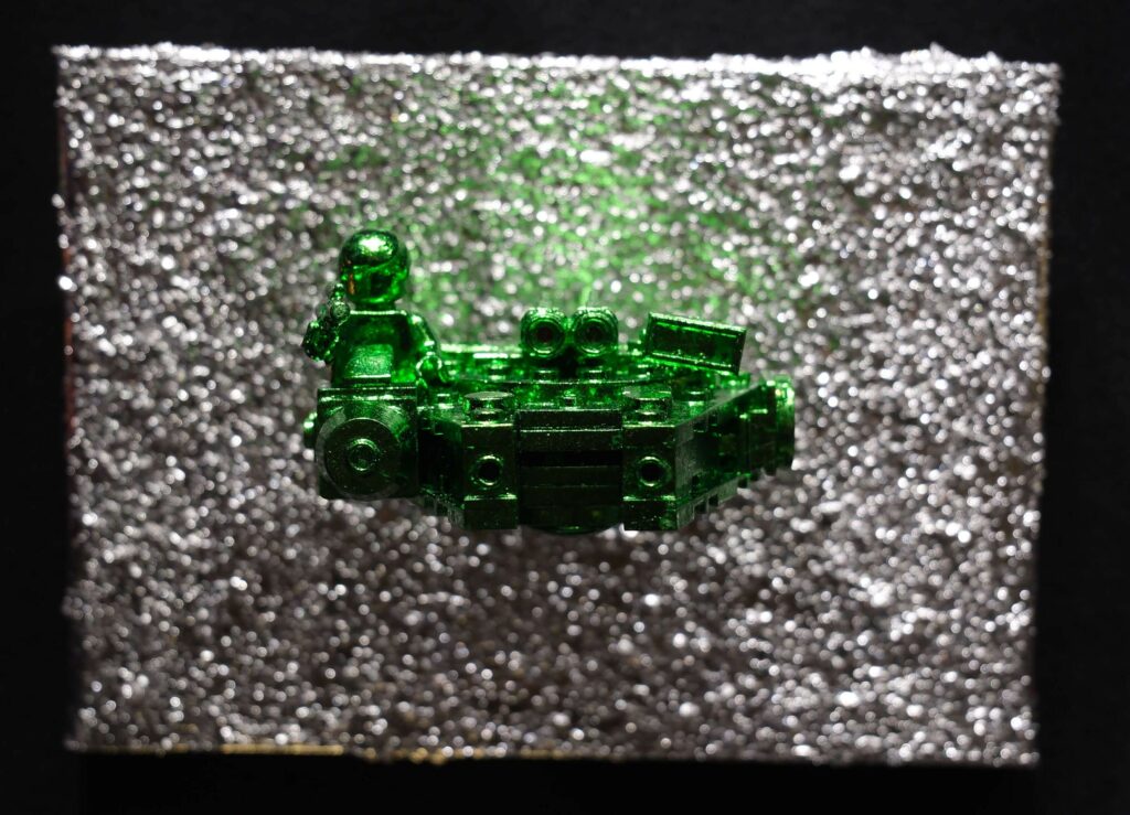 Seb Machten contemporary artwork with green starfighter built from construction bricks projecting from textured silver surface